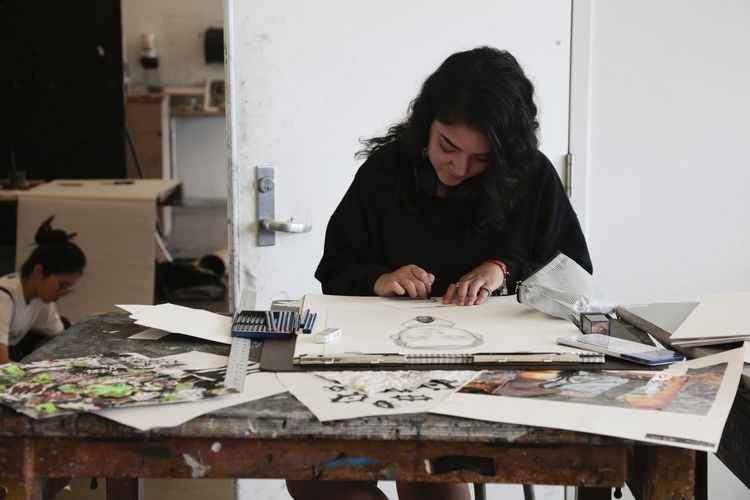 Student working on a drawing at a table covered in art supplies